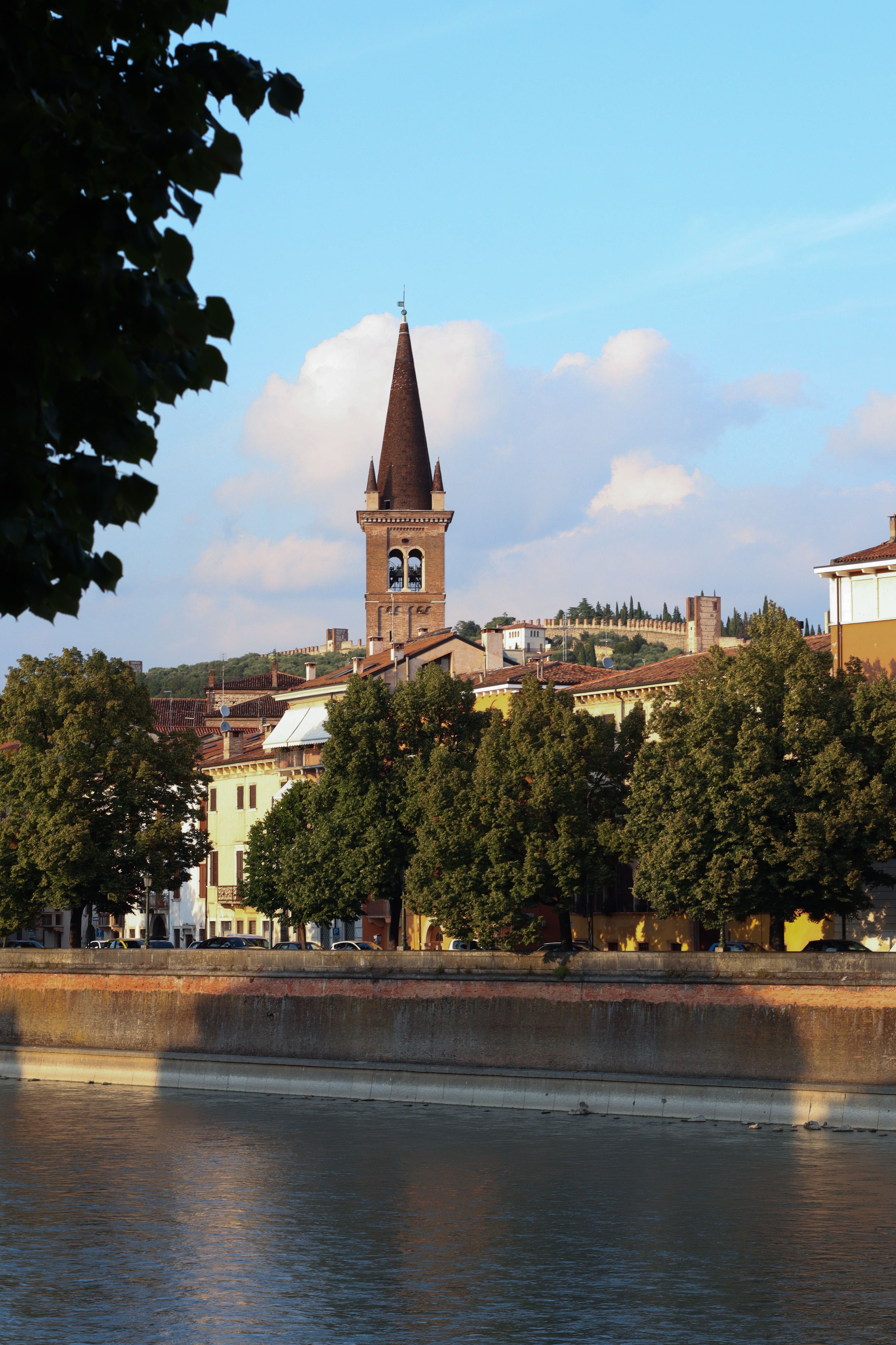 Picturesque view of a historic bell tower by the river, surrounded by lush trees and buildings.