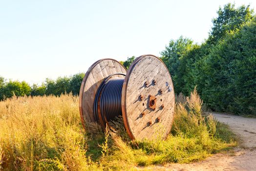 Wooden cable reel with coiled wires in a sunlit field. Ideal infrastructure stock photo.