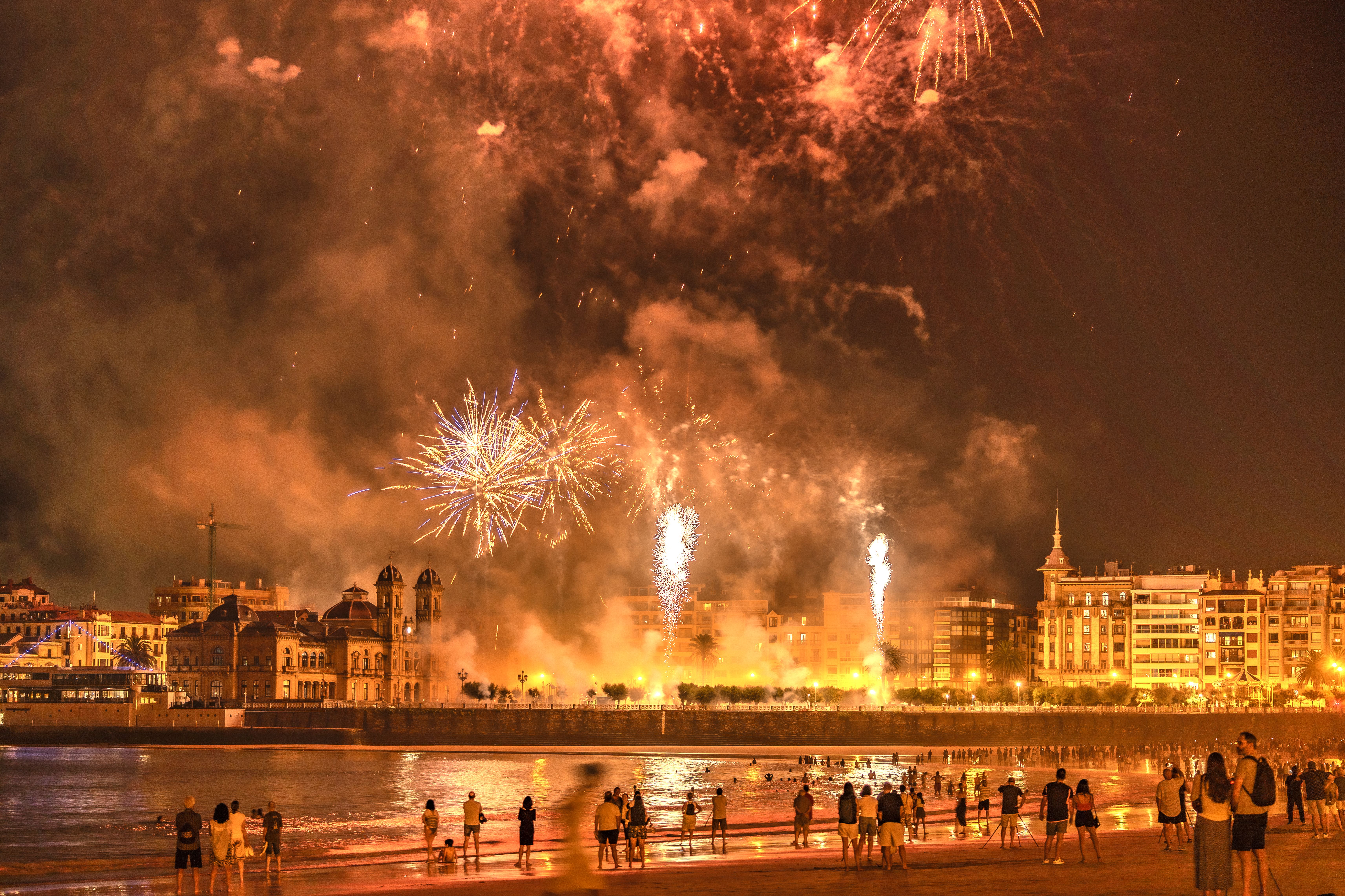 Nighttime fireworks lighting up the sky over a city beach with people watching. — Trójmiasto