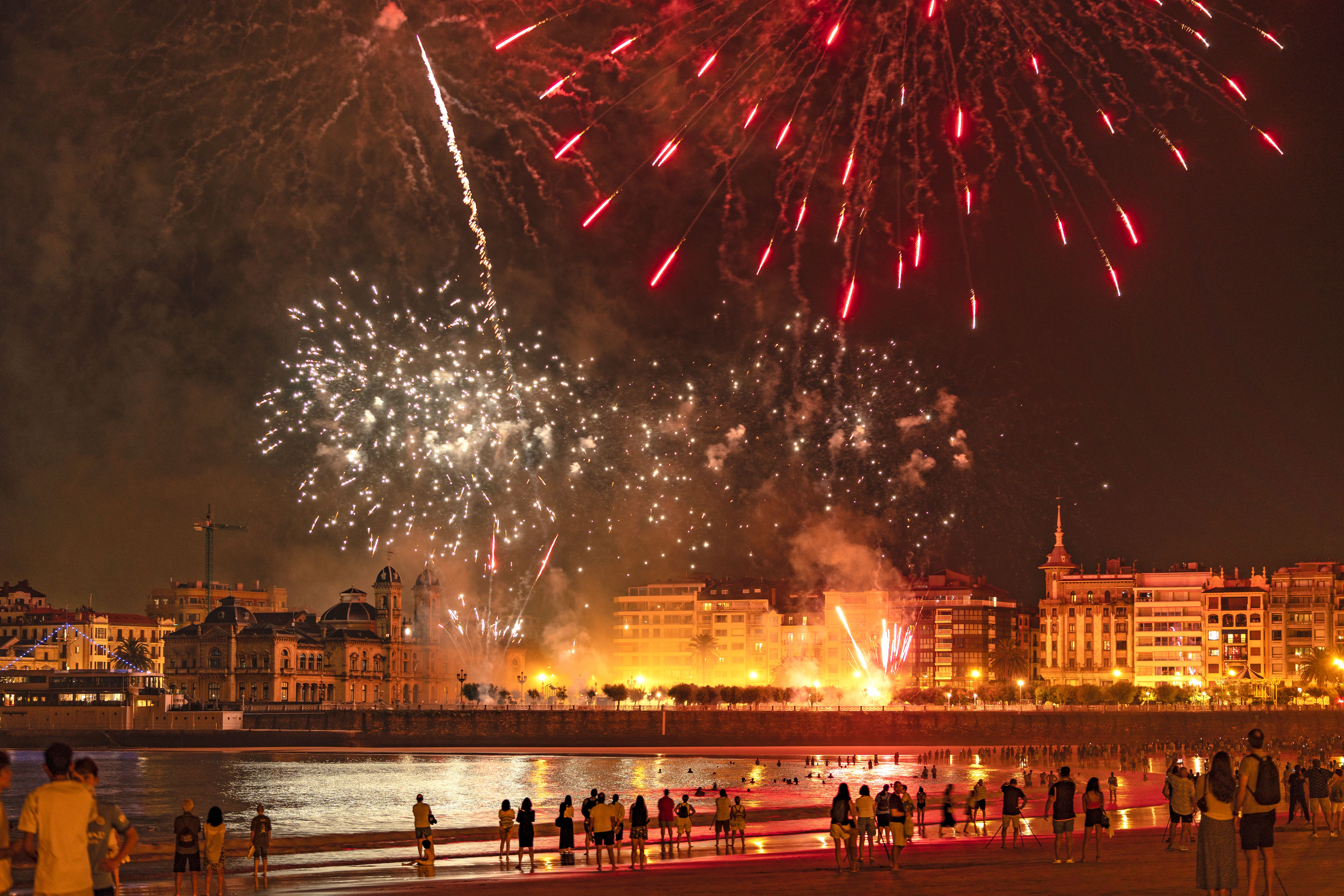 Des feux d'artifice spectaculaires illuminent le ciel nocturne au-dessus d'un front de mer animé, se reflétant sur l'eau en contrebas.