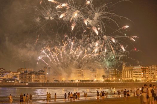 A vibrant fireworks show illuminates the coastal city at night with silhouettes on the beach.
