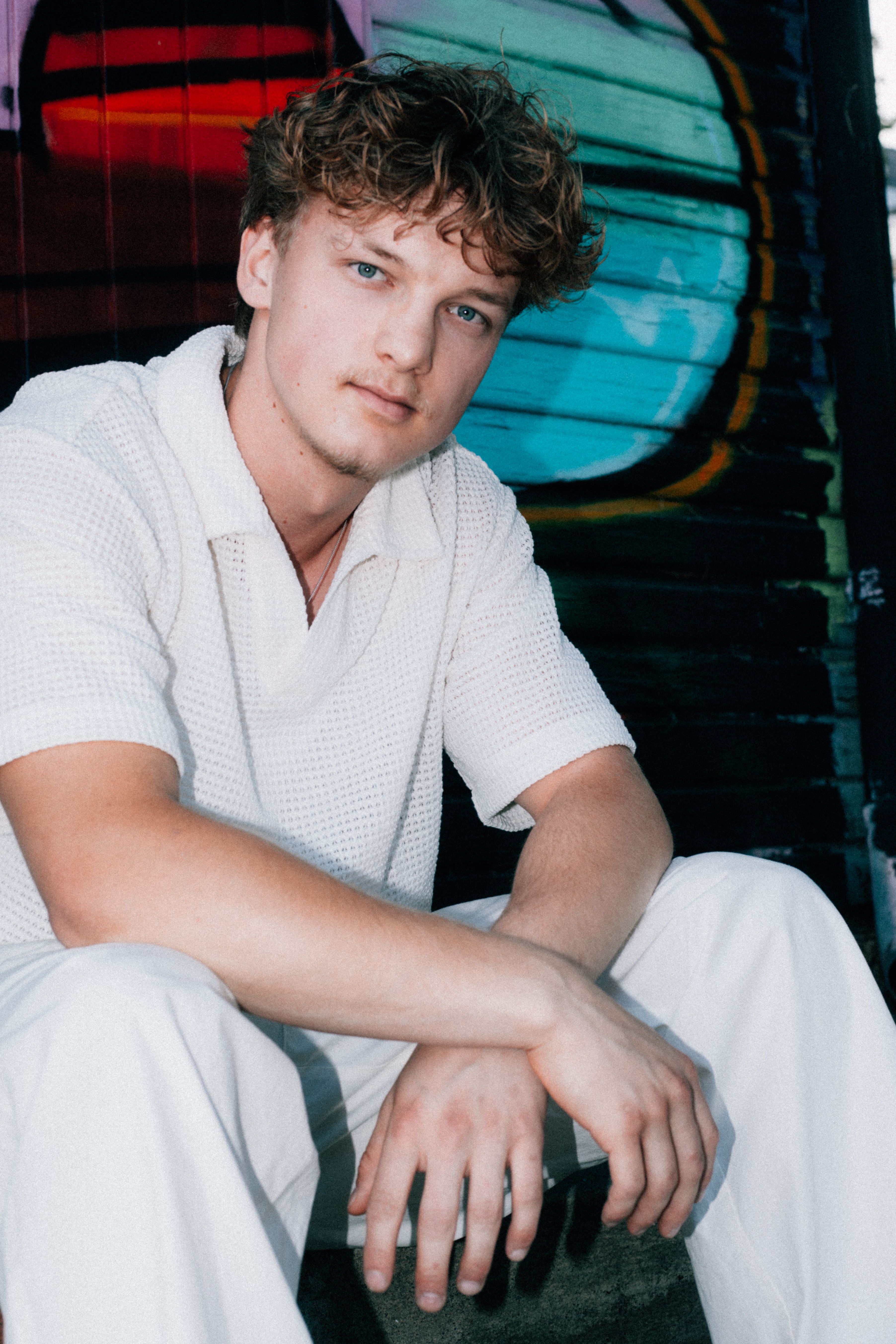 Portrait of a young man sitting near vibrant graffiti, wearing casual white attire, with a contemplative look.