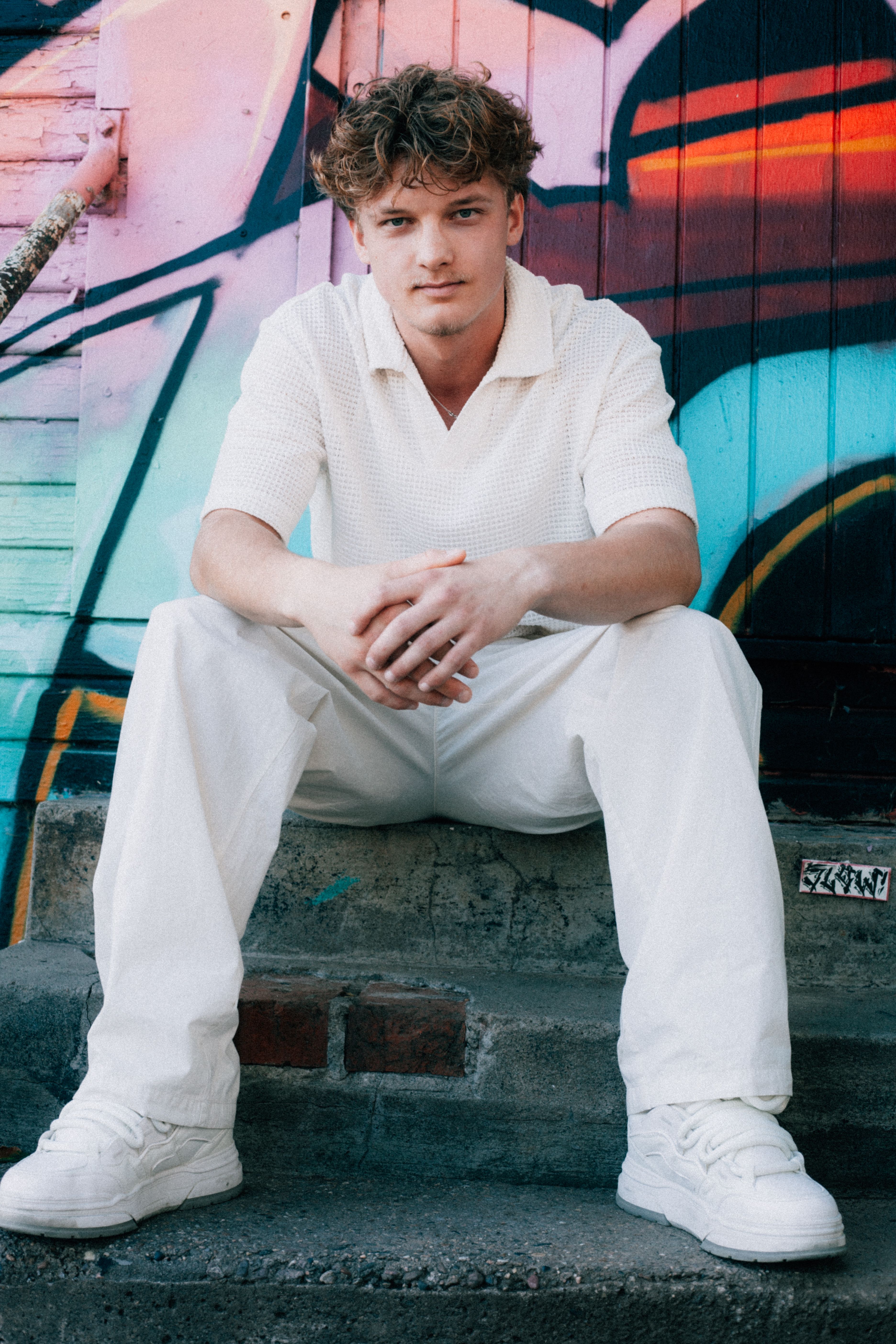 Casual portrait of a young man in white attire sitting on graffiti-decorated steps.