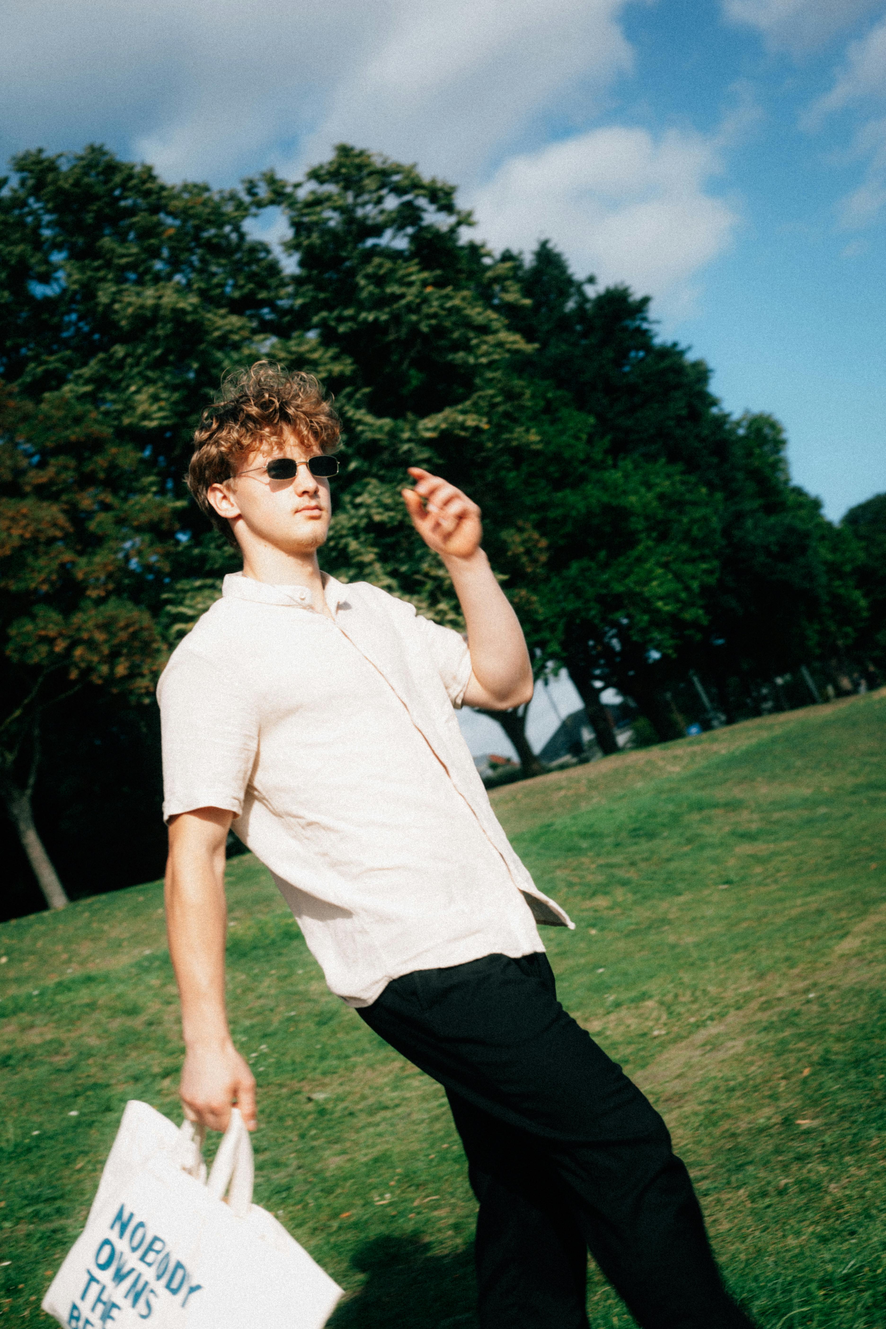 A man strolls through a sunny park carrying a tote bag with trees in the background.