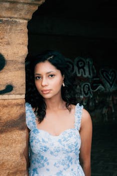 A young woman in a floral dress poses next to a graffiti-covered wall, exuding confidence and elegance.