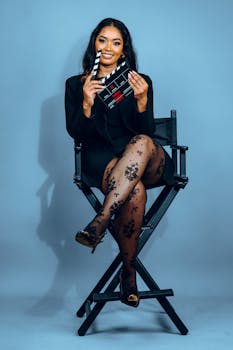 Elegant woman seated on director's chair holding clapperboard, dressed in black with patterned stockings.