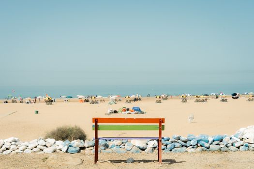 A vibrant bench facing a lively beach filled with umbrellas under a clear sky.