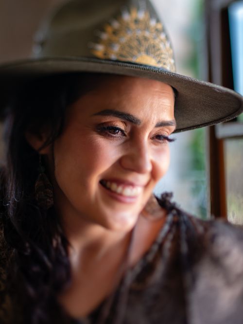 Free Portrait of a smiling woman wearing a stylish hat by a window in warm lighting. Stock Photo