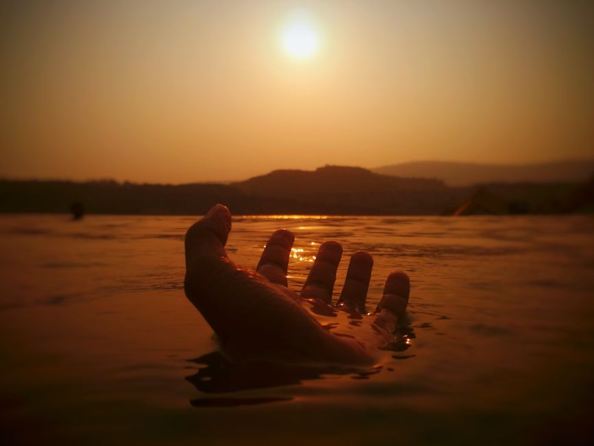 A dramatic scene of a hand reaching towards the sunset over a calm body of water, conveying a sense of serenity.