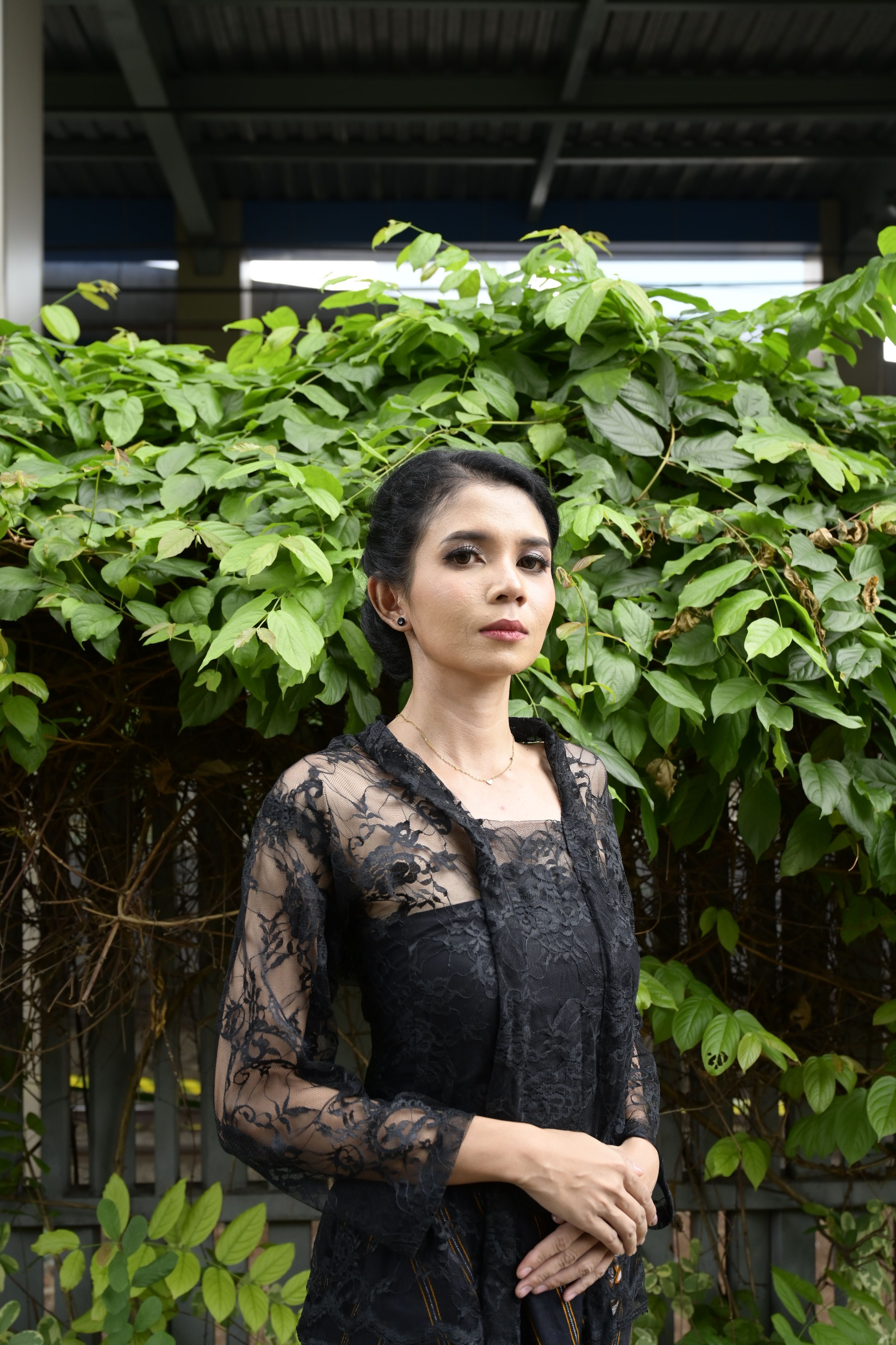 A woman in a black lace kebaya poses outdoors in Jakarta. Lush greenery backdrop.
