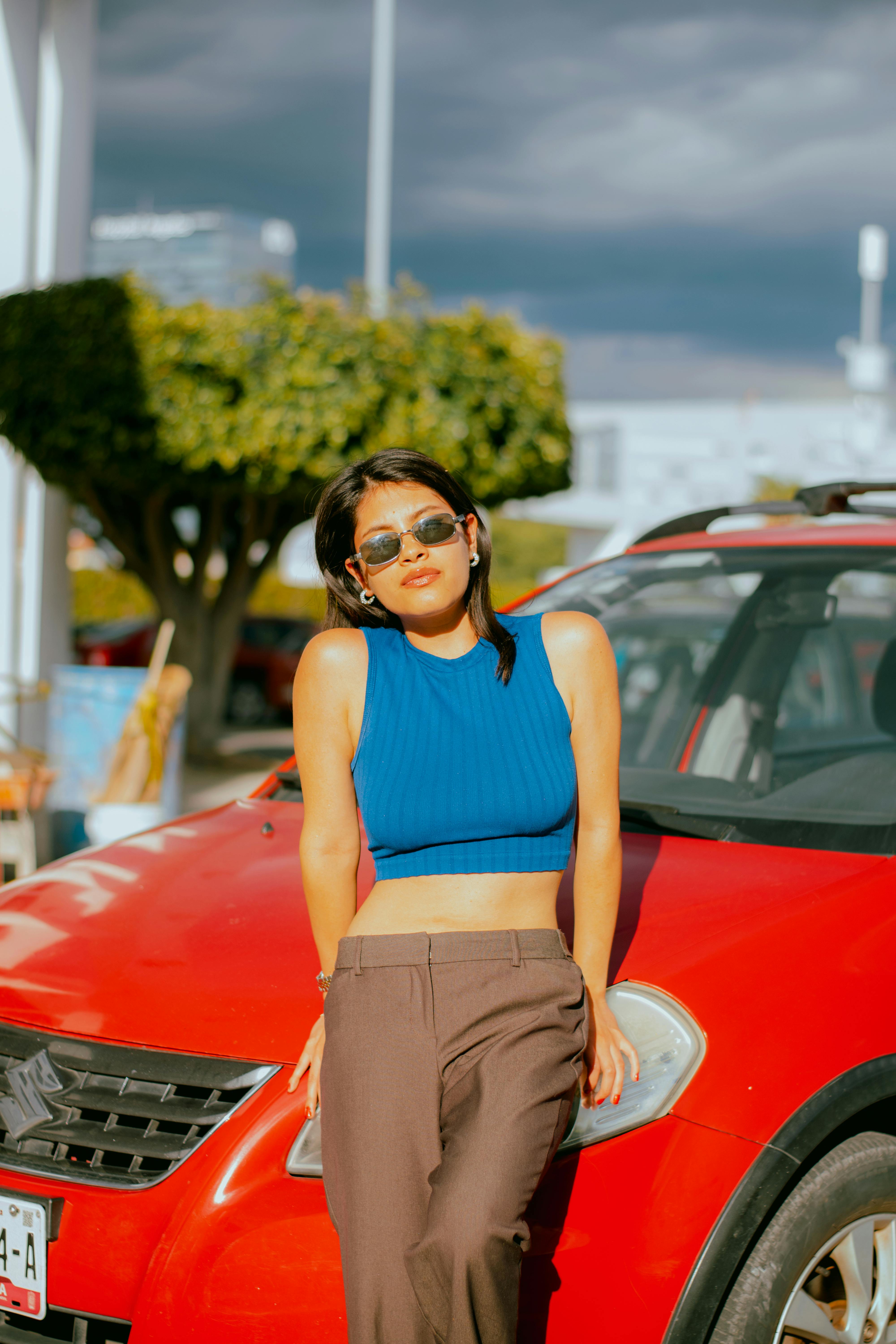 Free Young woman in sunglasses posing confidently by red car under a sunny sky. Stock Photo