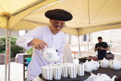 A street vendor in traditional attire pours tea into cups at an outdoor market under a yellow tent.