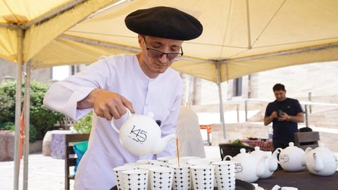 A street vendor in traditional attire pours tea into cups at an outdoor market under a yellow tent.