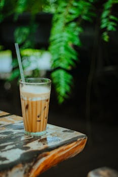 Refreshing iced coffee in glass with straw on rustic table outdoors, surrounded by greenery.