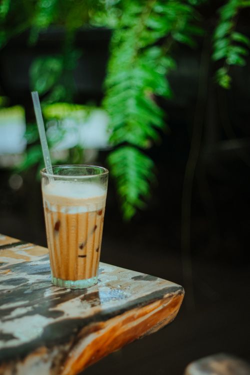 Free Refreshing iced coffee in glass with straw on rustic table outdoors, surrounded by greenery. Stock Photo