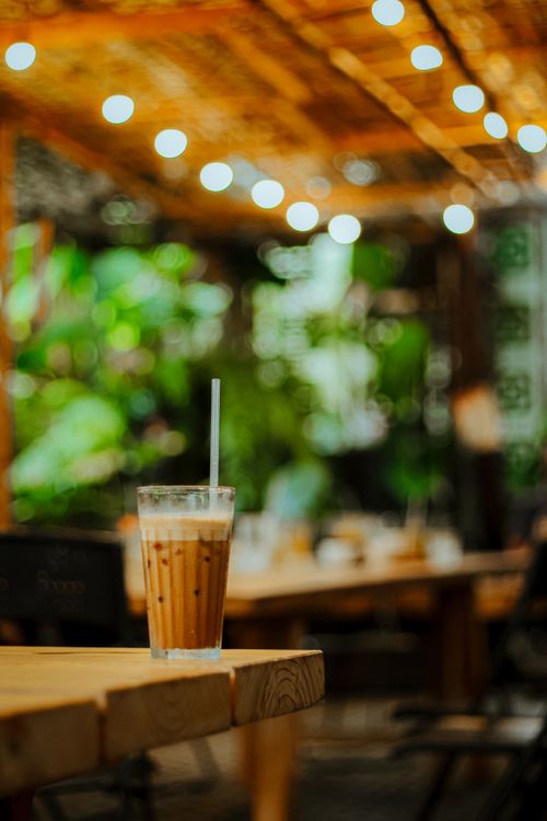 Free A refreshing glass of iced coffee on a wooden table in a cozy outdoor café with string lights. Stock Photo