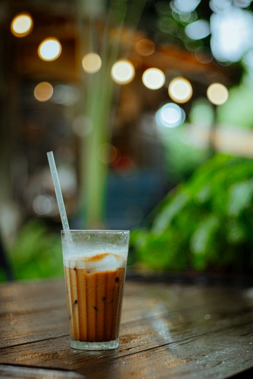 Free A glass of iced coffee with straw on a wet wooden table, outdoors with bokeh lights. Stock Photo
