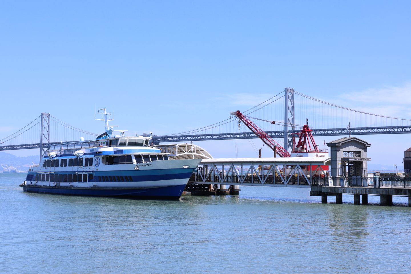 A San Francisco ferry docked at a pier with the Bay Bridge in the background on a clear sunny day