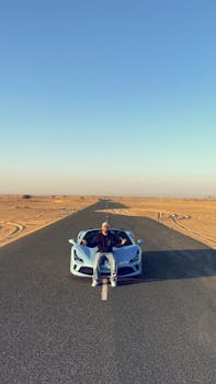 A man sits on a luxury sports car parked on a desert highway under a clear blue sky.
