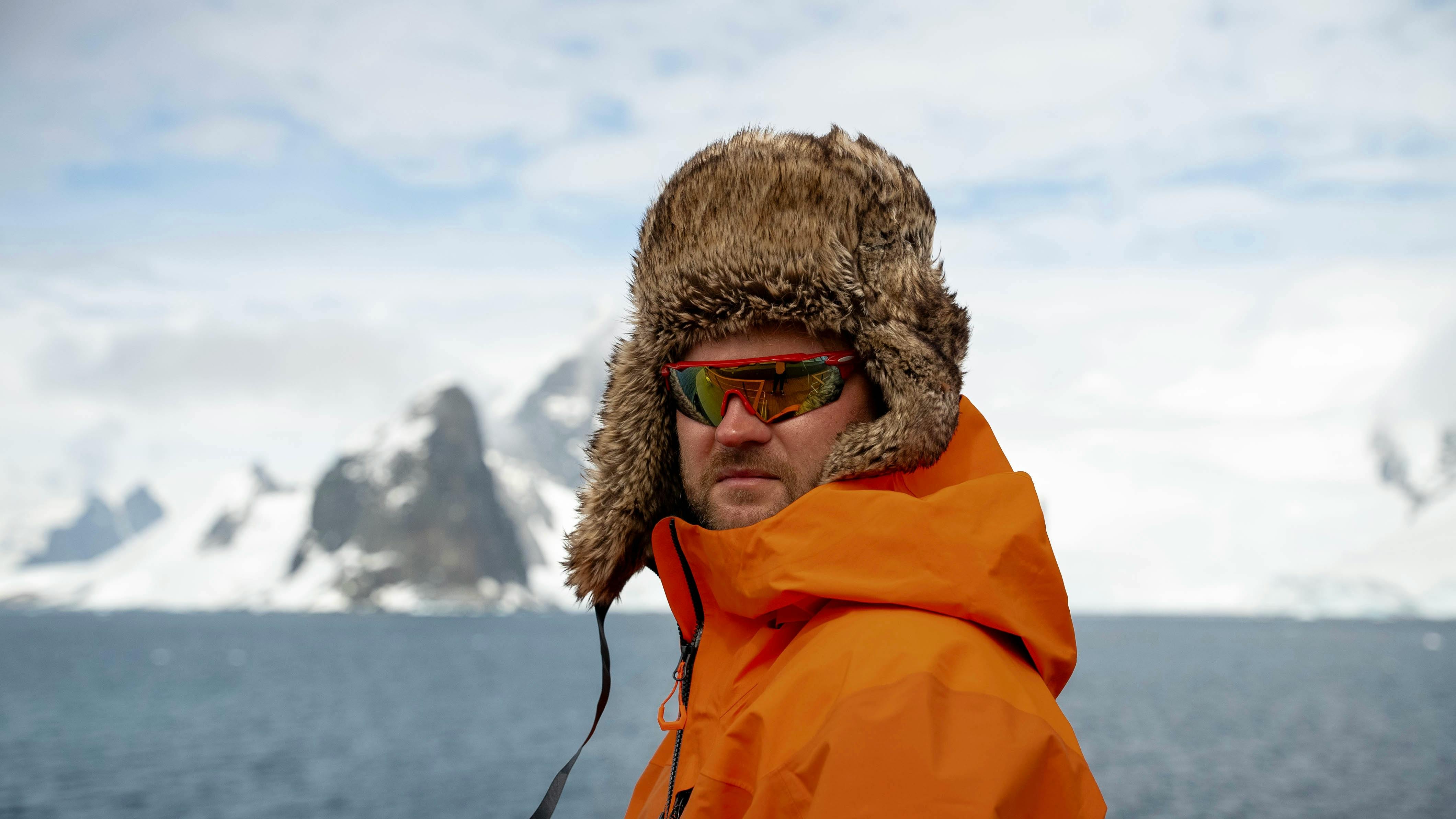 Man in orange parka and fur hat exploring Antarctica with icy mountains in the background.