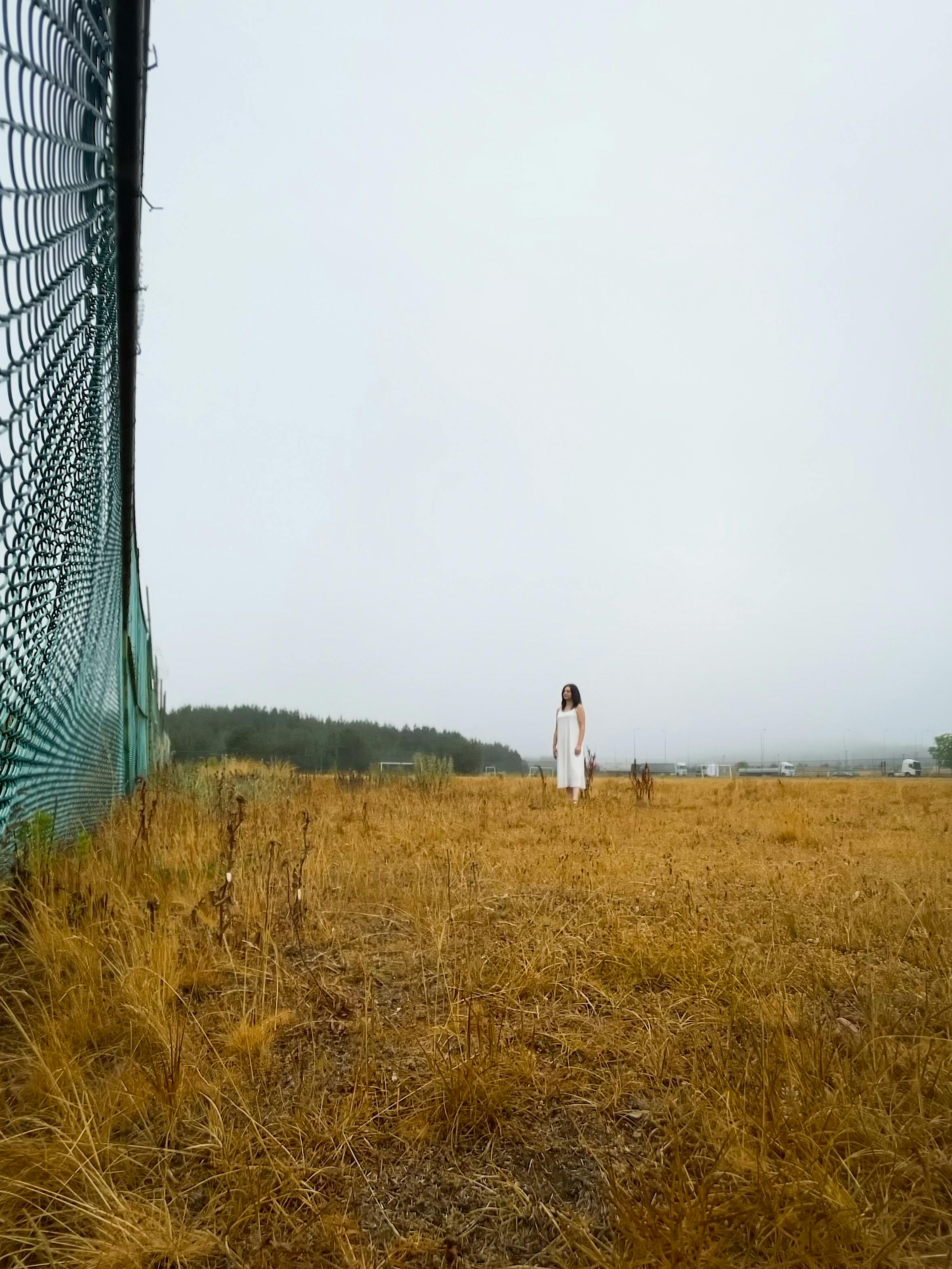 Gratis En kvinde i en hvid kjole står alene på en åben mark nær et hegn og skaber en rolig og isoleret atmosfære. Lagerfoto