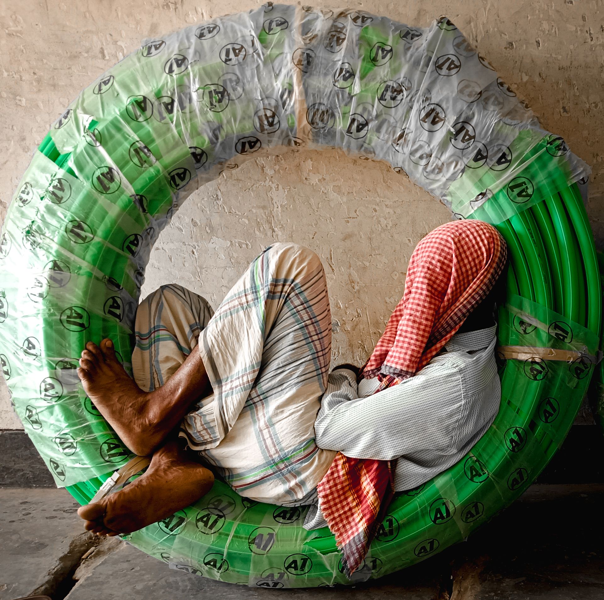 A man in traditional attire rests inside a large coiled green pipe against a wall.