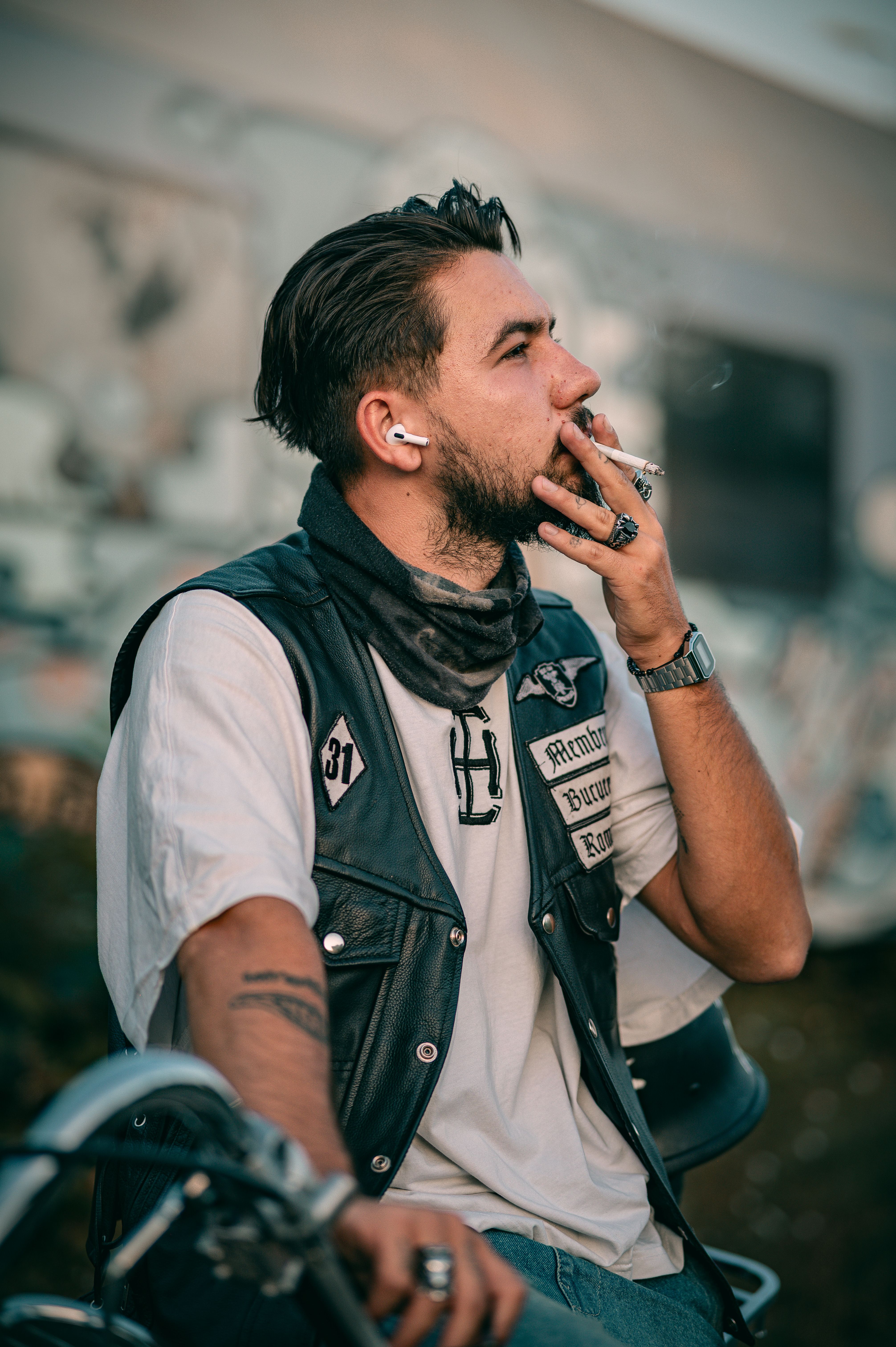 Stylish young biker in a leather vest smokes a cigarette in an urban background.
