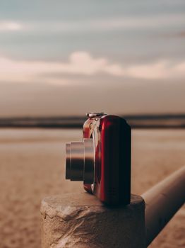 A red vintage camera with metal lens on a beach rail at sunset, contrasting with a blurred background.