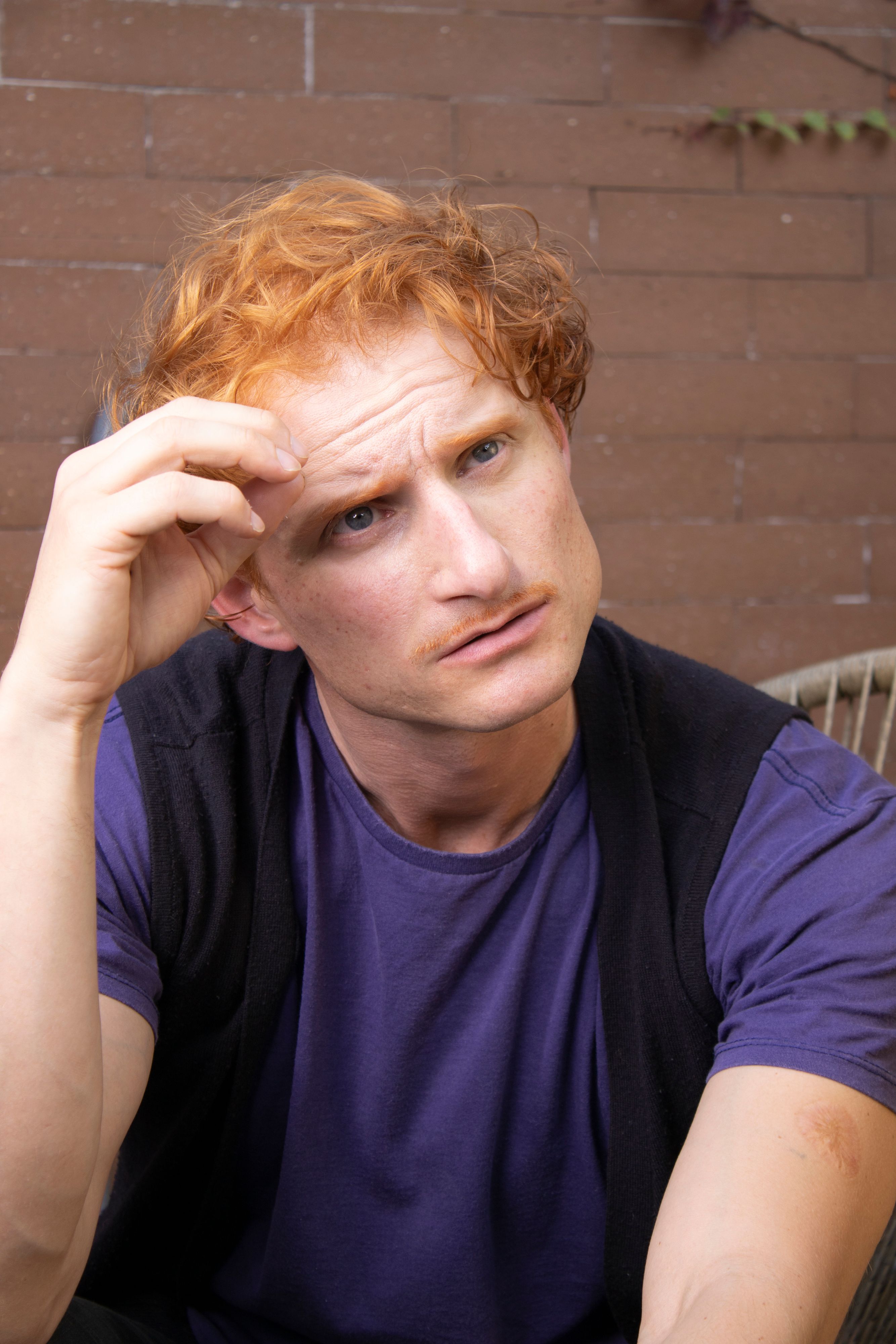 A thoughtful man with red hair in casual attire poses outdoors on a sunny day.