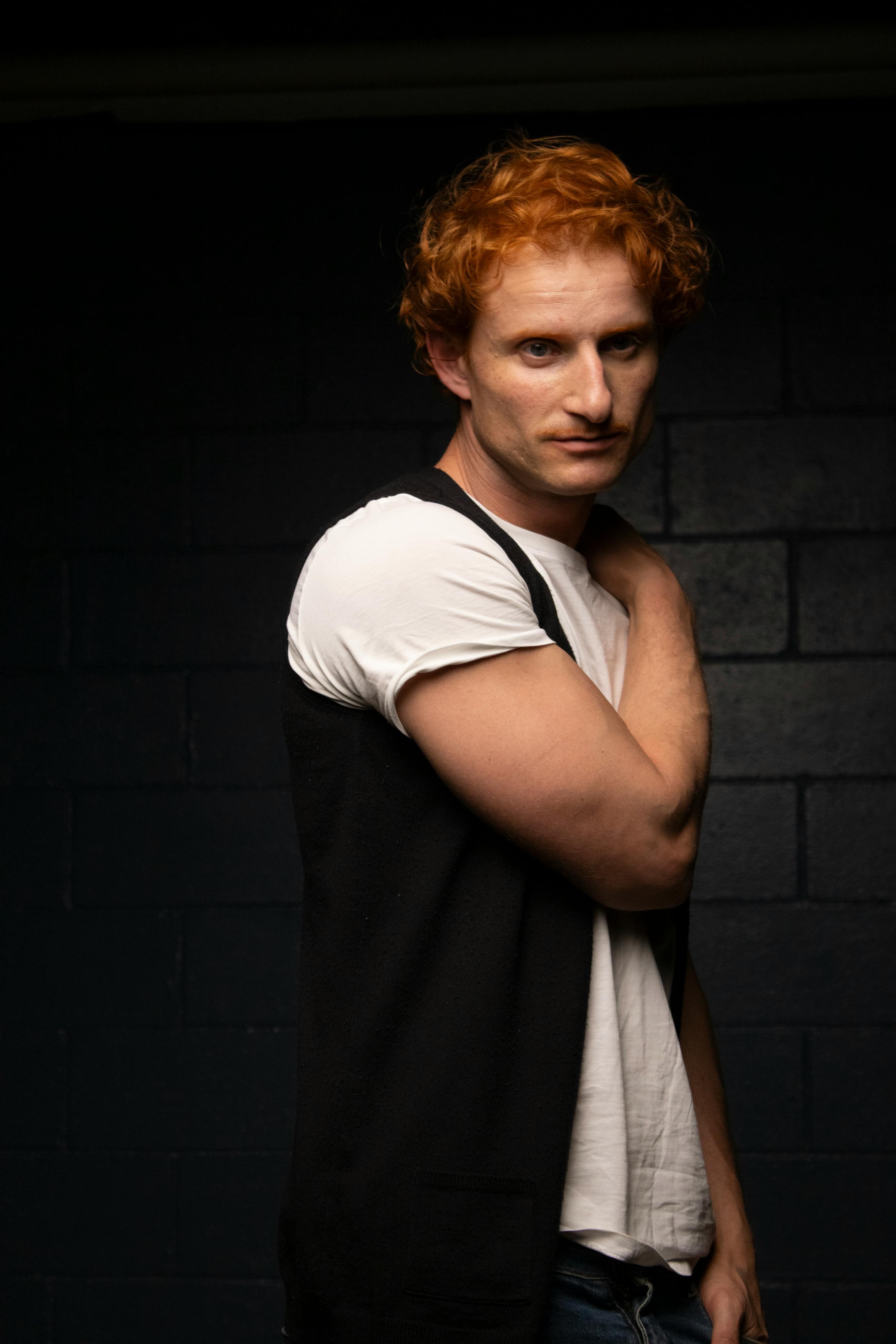 A contemplative red-haired man poses in casual attire against a dark background.