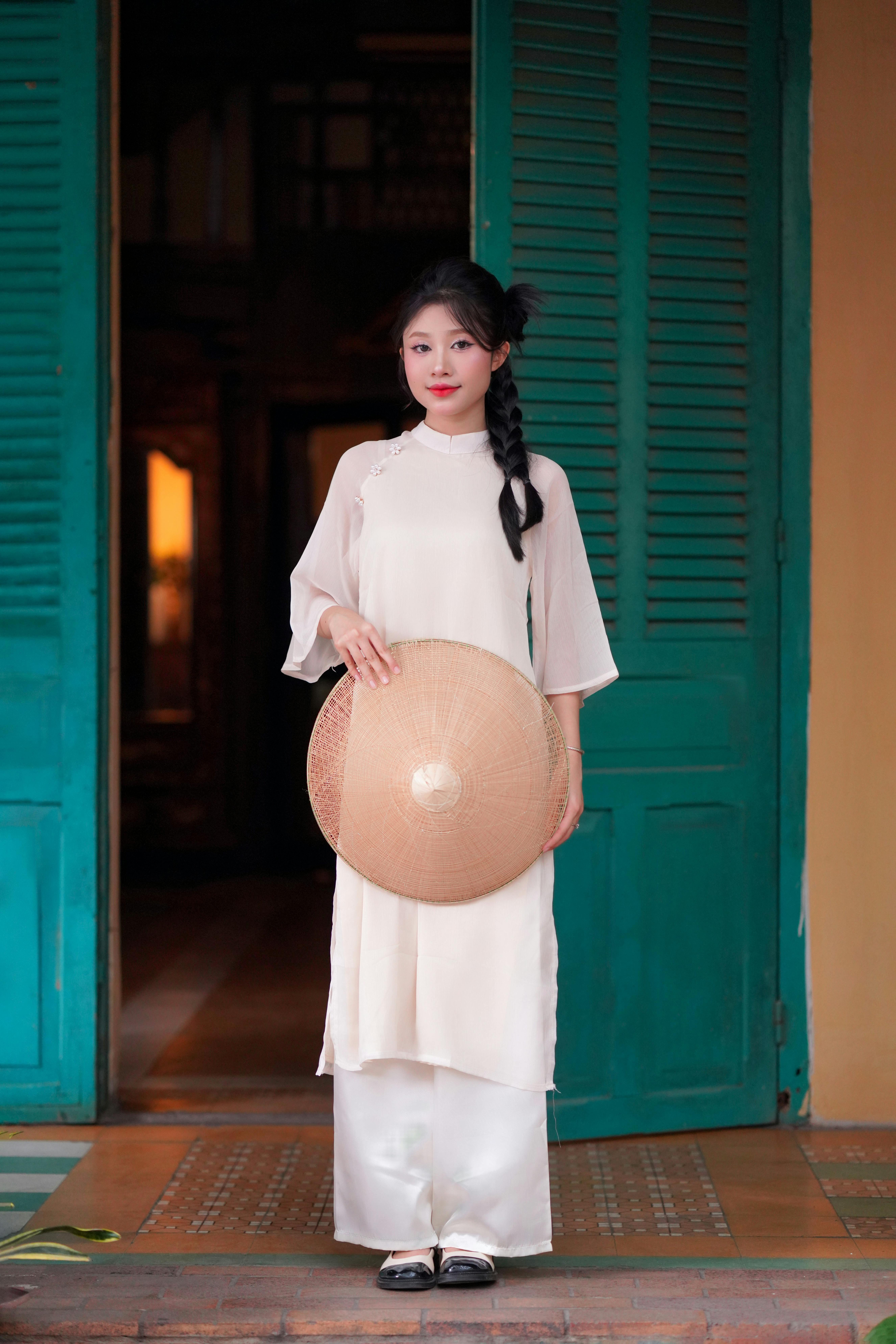 Portrait of a woman in traditional Vietnamese attire holding a conical hat, standing by blue shutters.