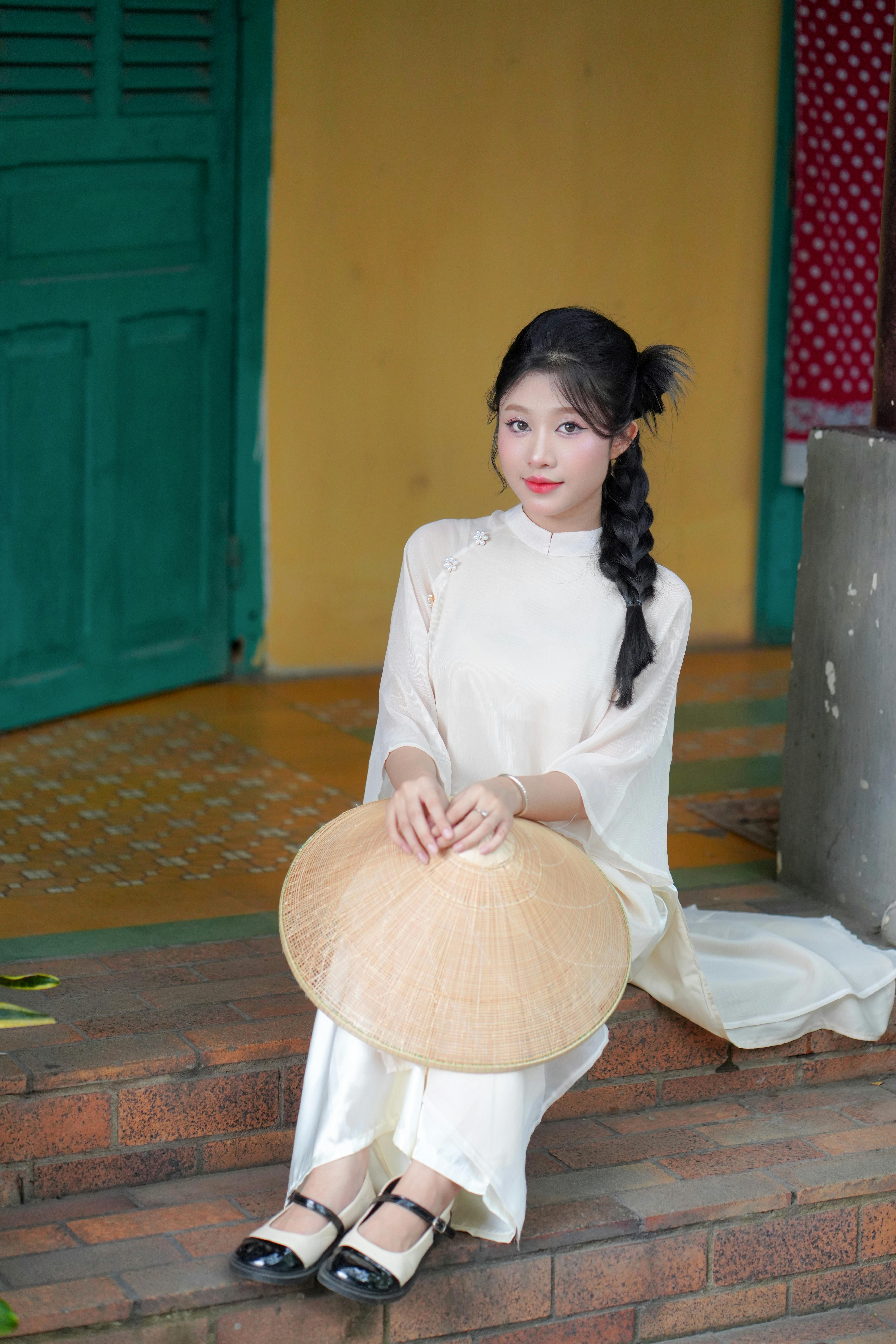 A young woman in a traditional Ao Dai holding a conical hat, sitting on steps.