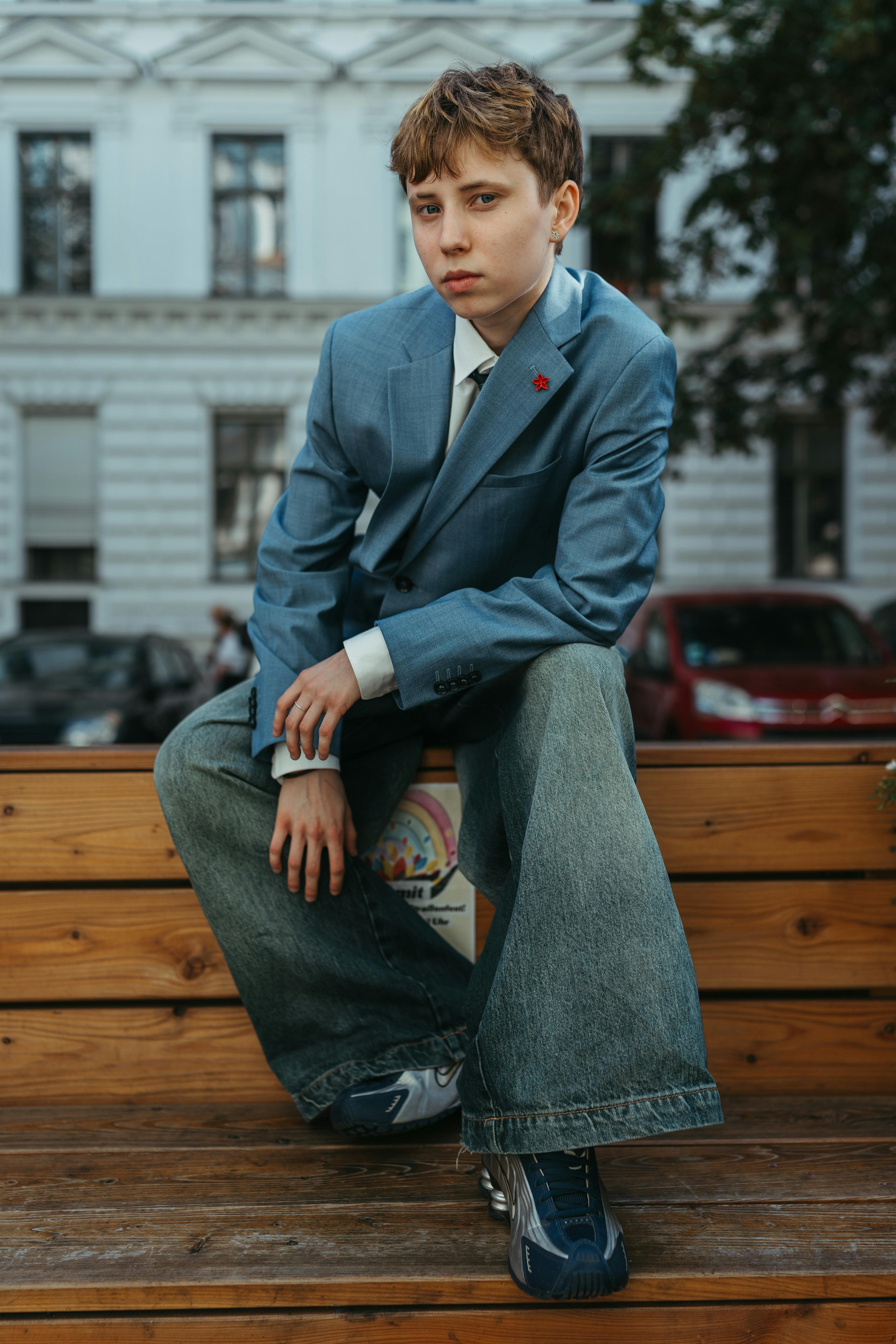 A fashionable teenager poses in denim jeans and a blue blazer on a wooden bench in urban Berlin.
