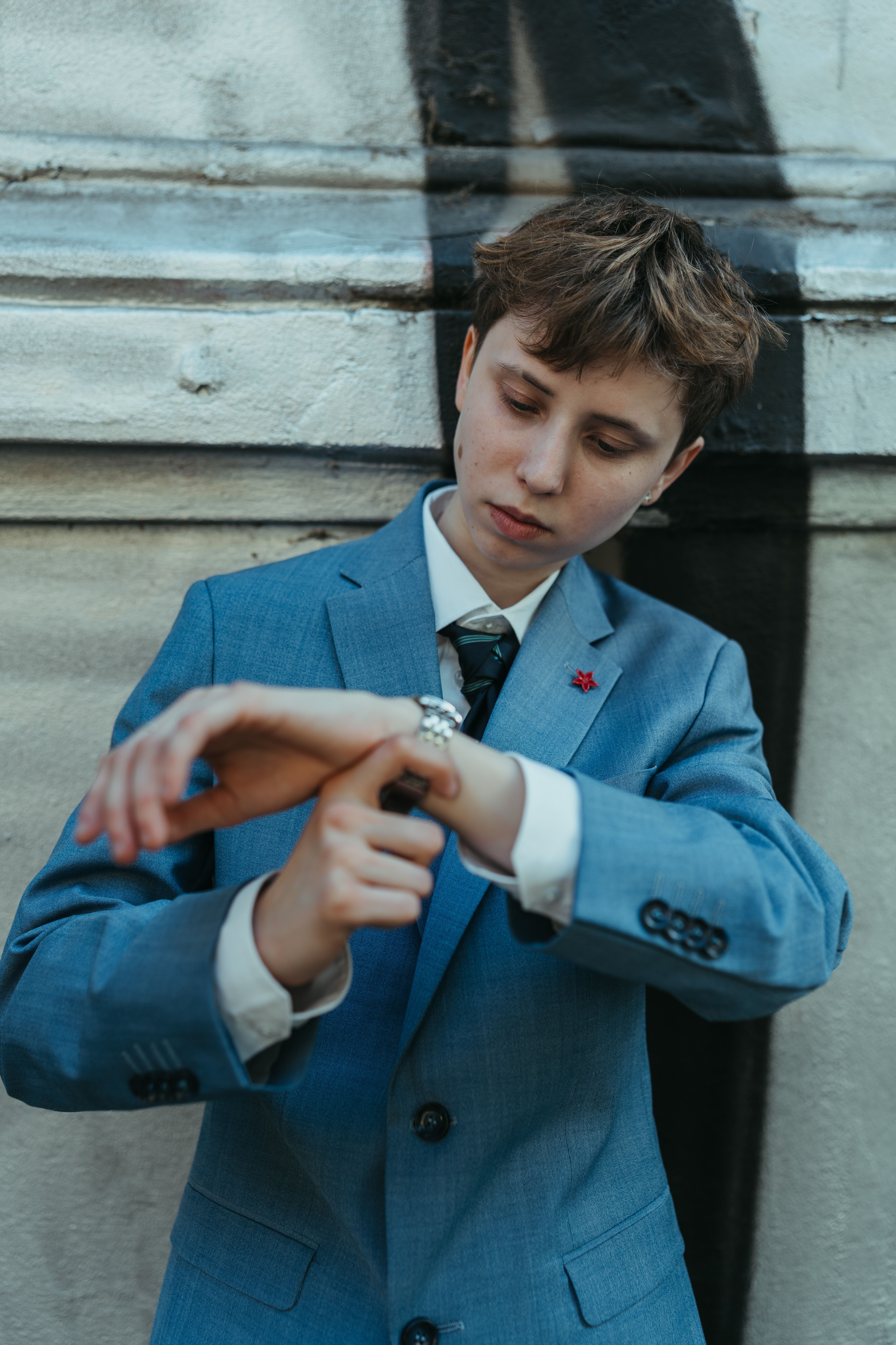 A young adult in a blue suit checks their watch against a city wall with urban graffiti.