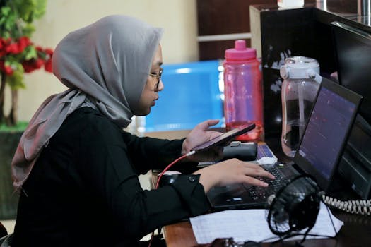 Young woman using phone and laptop in an office setting, multitasking and focusing on work.