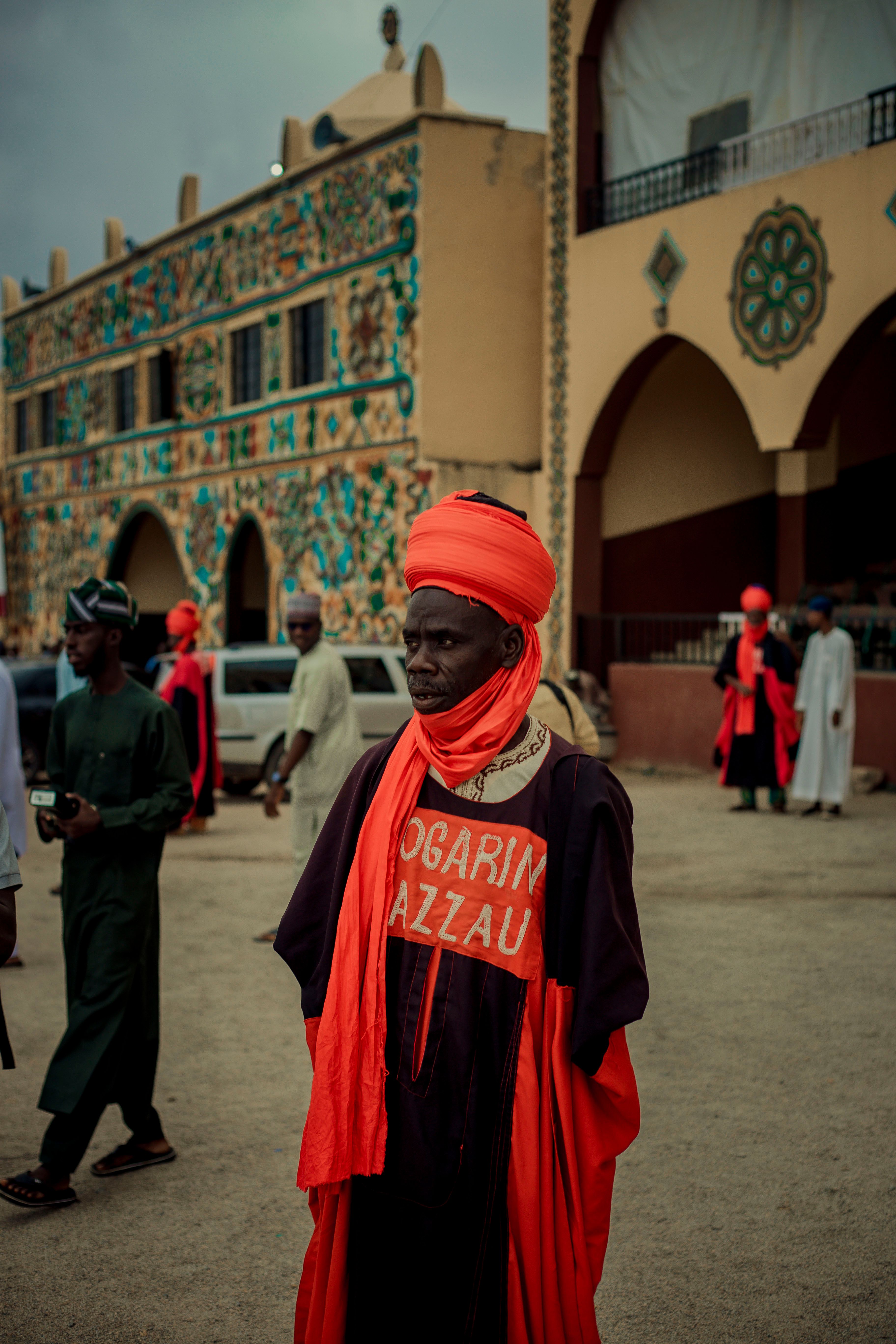 Man in traditional Nigerian attire with vibrant colors stands in a decorated street setting, showcasing cultural richness.