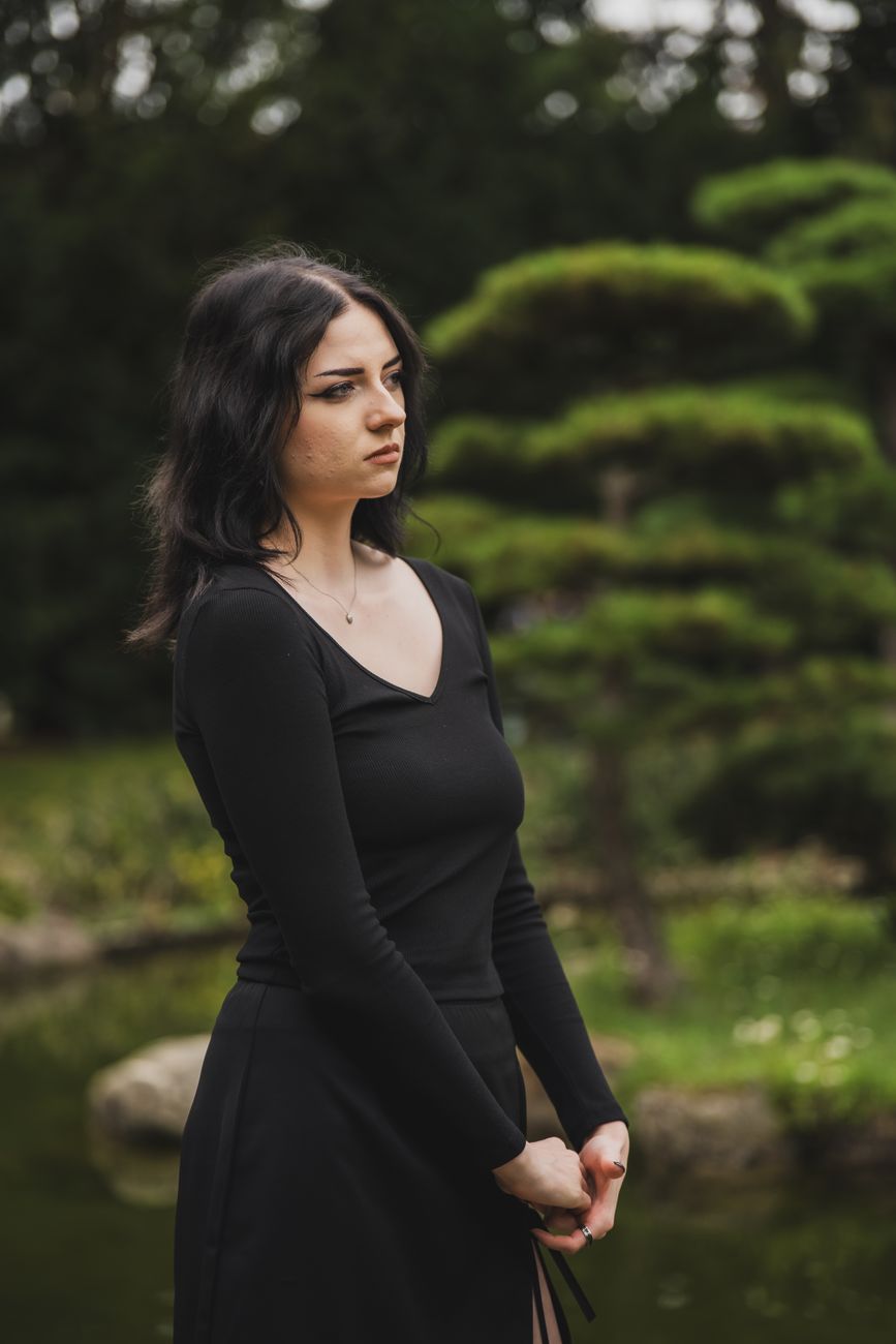 A woman in black attire standing thoughtfully in a tranquil Japanese garden setting.