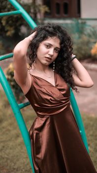 Portrait of a curly-haired woman in a stylish satin dress posing gracefully in an outdoor setting.