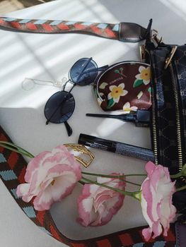 Stylish flat lay of sunglasses, floral pouch, and makeup on a sunlit surface.