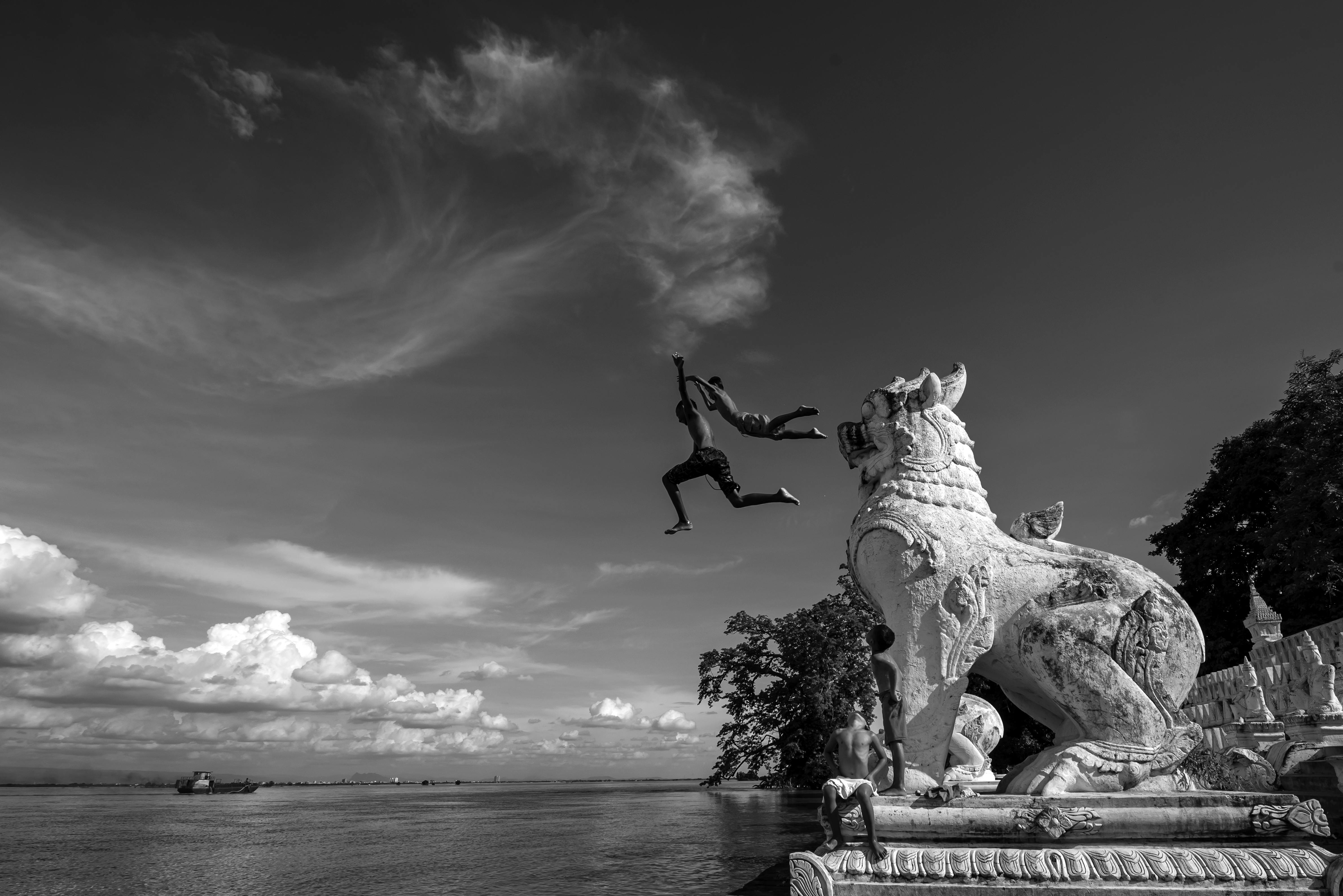 Captivating black and white scene of boys leaping by a mythical lion statue near a river in Min Kun, Myanmar.
