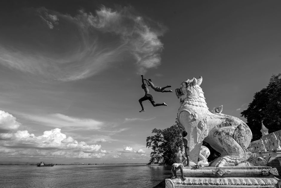Captivating black and white scene of boys leaping by a mythical lion statue near a river in Min Kun, Myanmar.