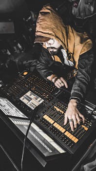 Hooded musician adjusting equipment in a modern recording studio control room.