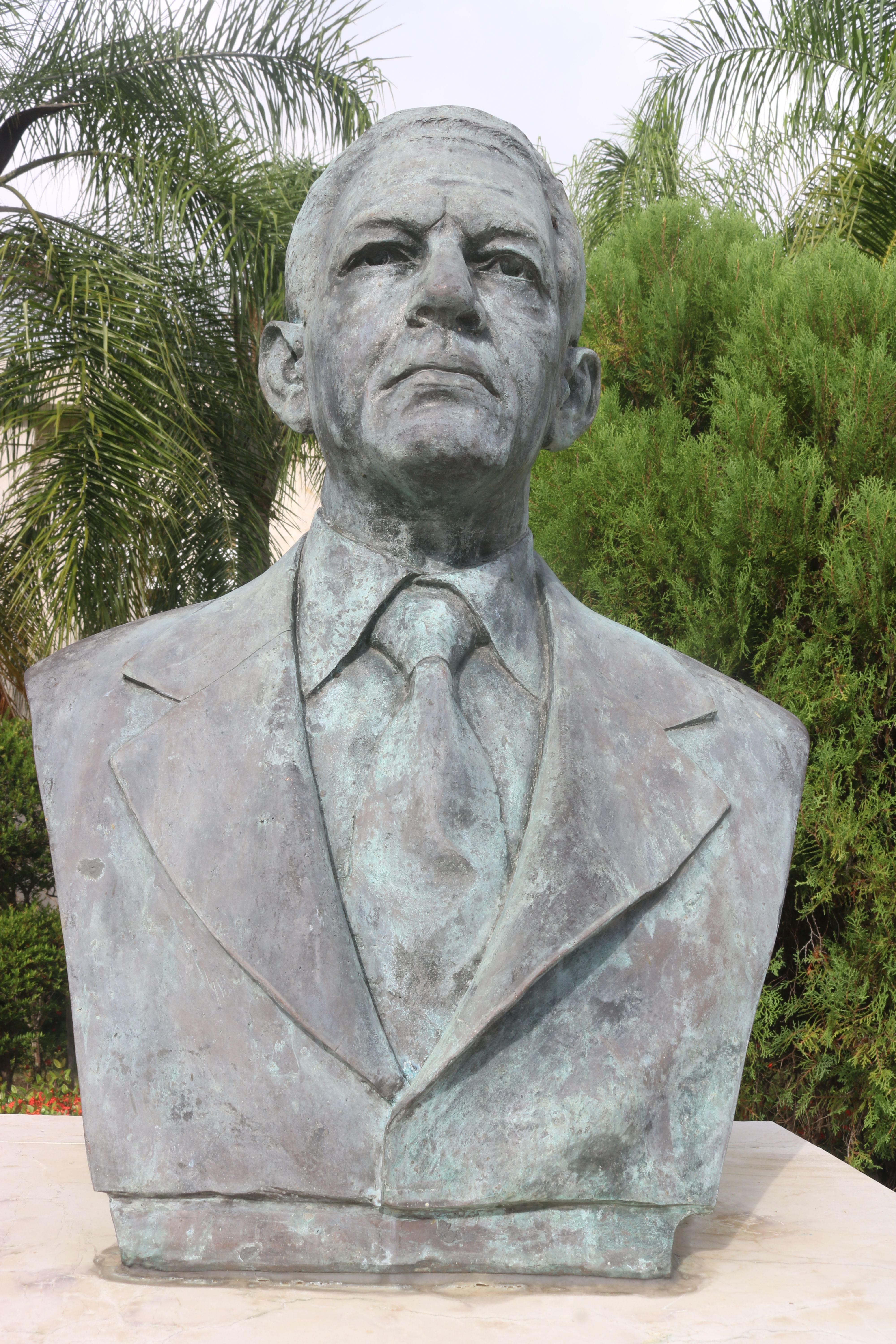 Bronze sculpture of a man in a suit, set outdoors against a lush green backdrop.