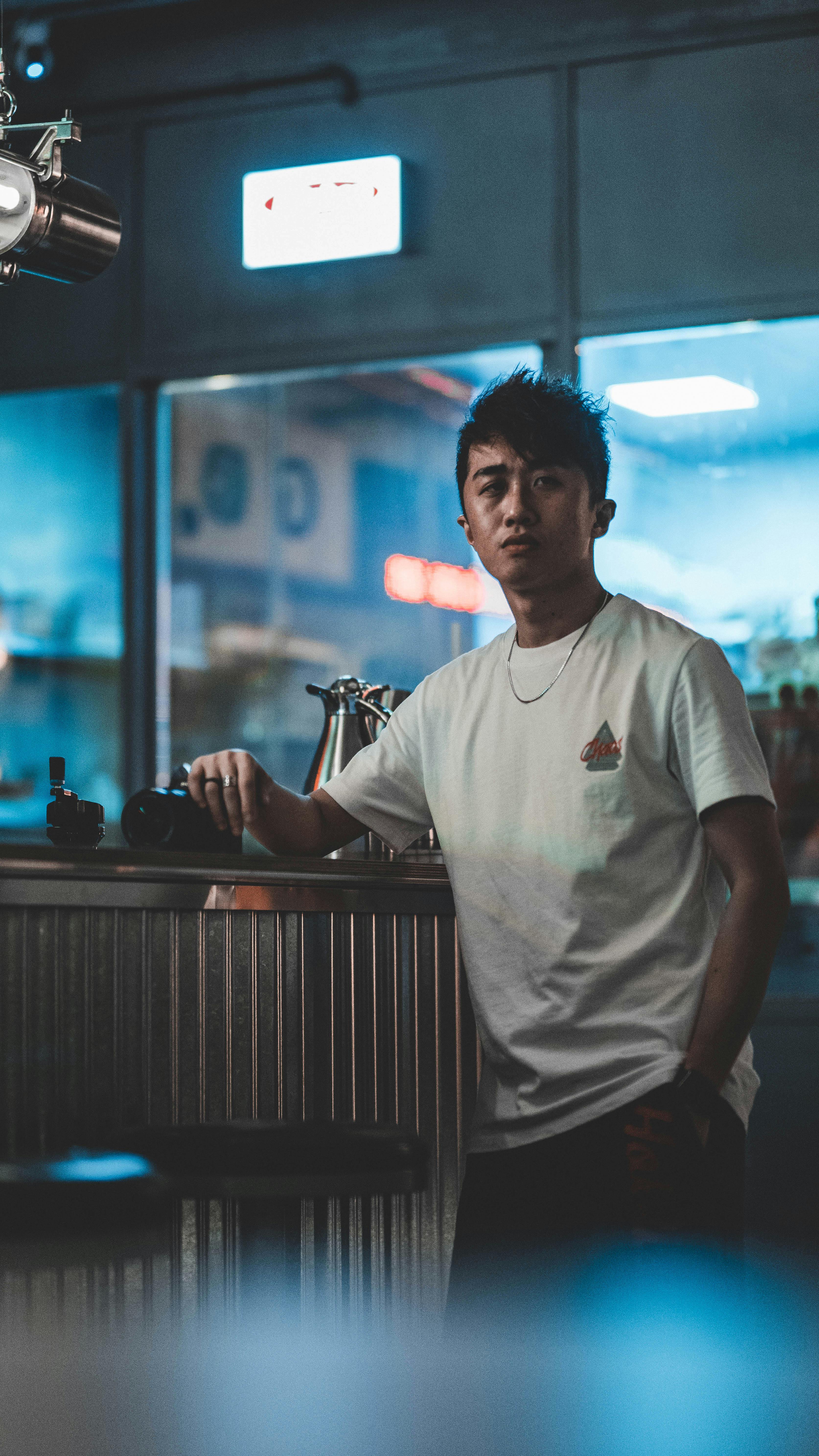 A young man in a white shirt standing at a counter in a modern cafe with moody lighting.