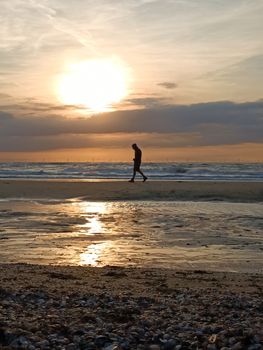 A serene silhouette of a person walking along a beach during a stunning sunset with reflections on wet sand.