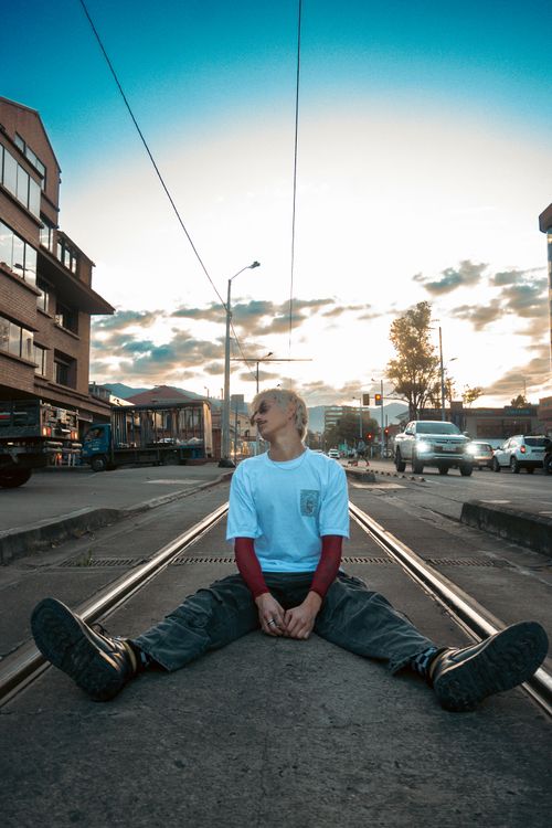 Free A man sits confidently on tram tracks in Cuenca, Ecuador, with a vibrant evening sky and urban backdrop. Stock Photo