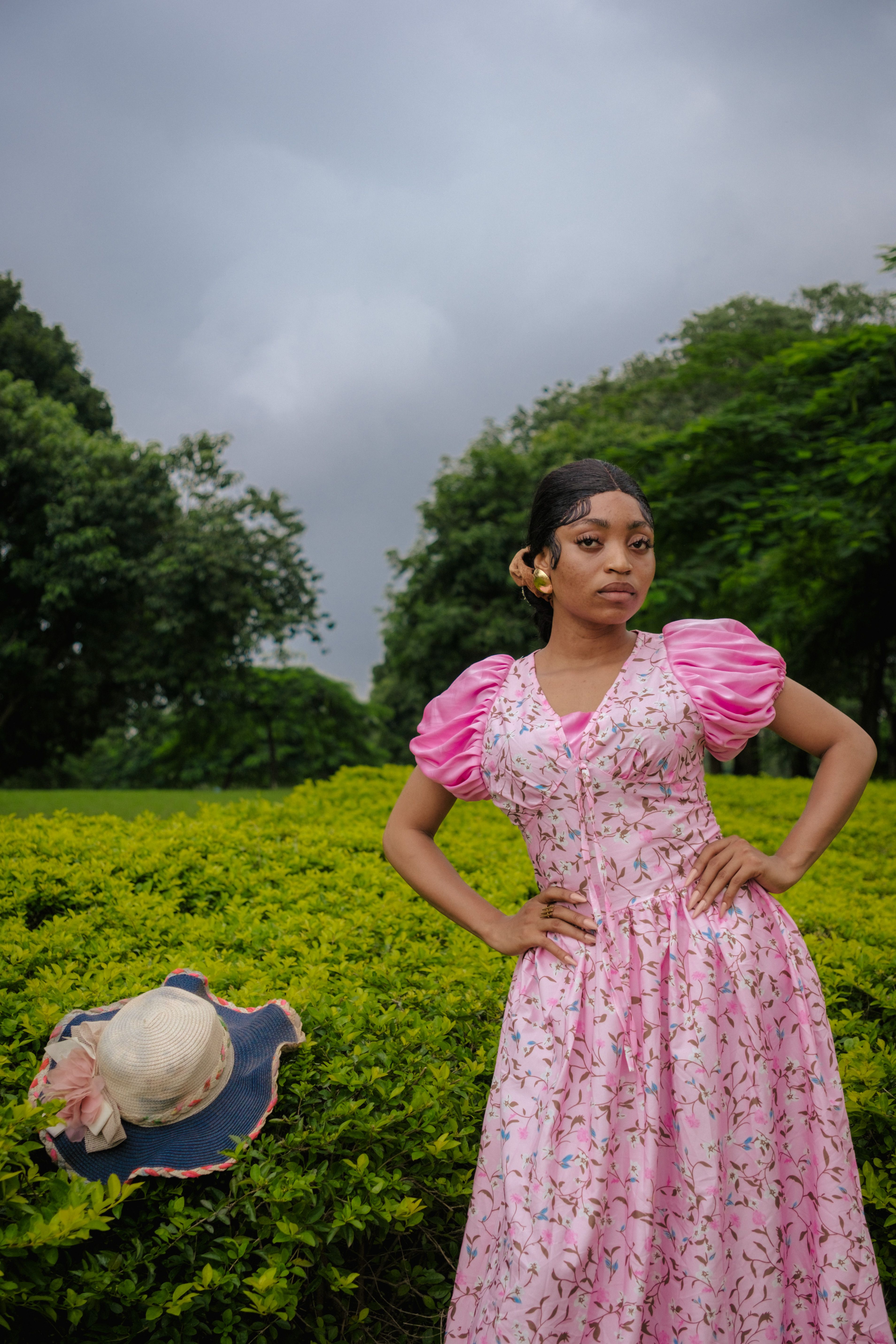 A woman in a pink floral dress poses confidently in a lush green garden under a cloudy sky.