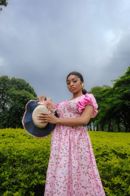 Free A woman in a floral pink dress holding a wide-brimmed hat in a lush green park. Stock Photo