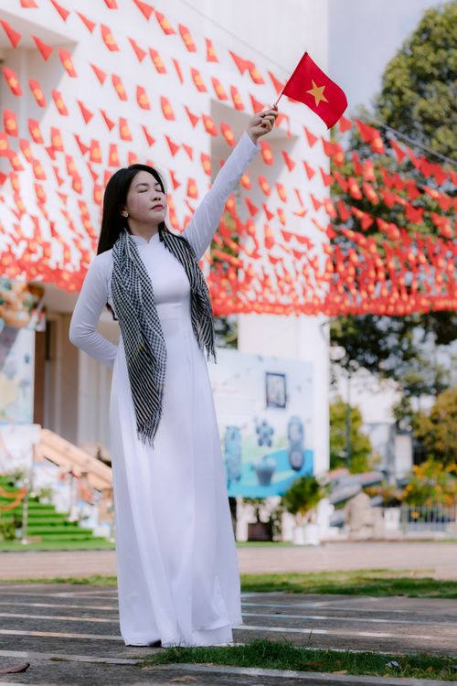 Free Woman in white traditional dress and scarf holding a red flag in front of decorated outdoor backdrop. Stock Photo