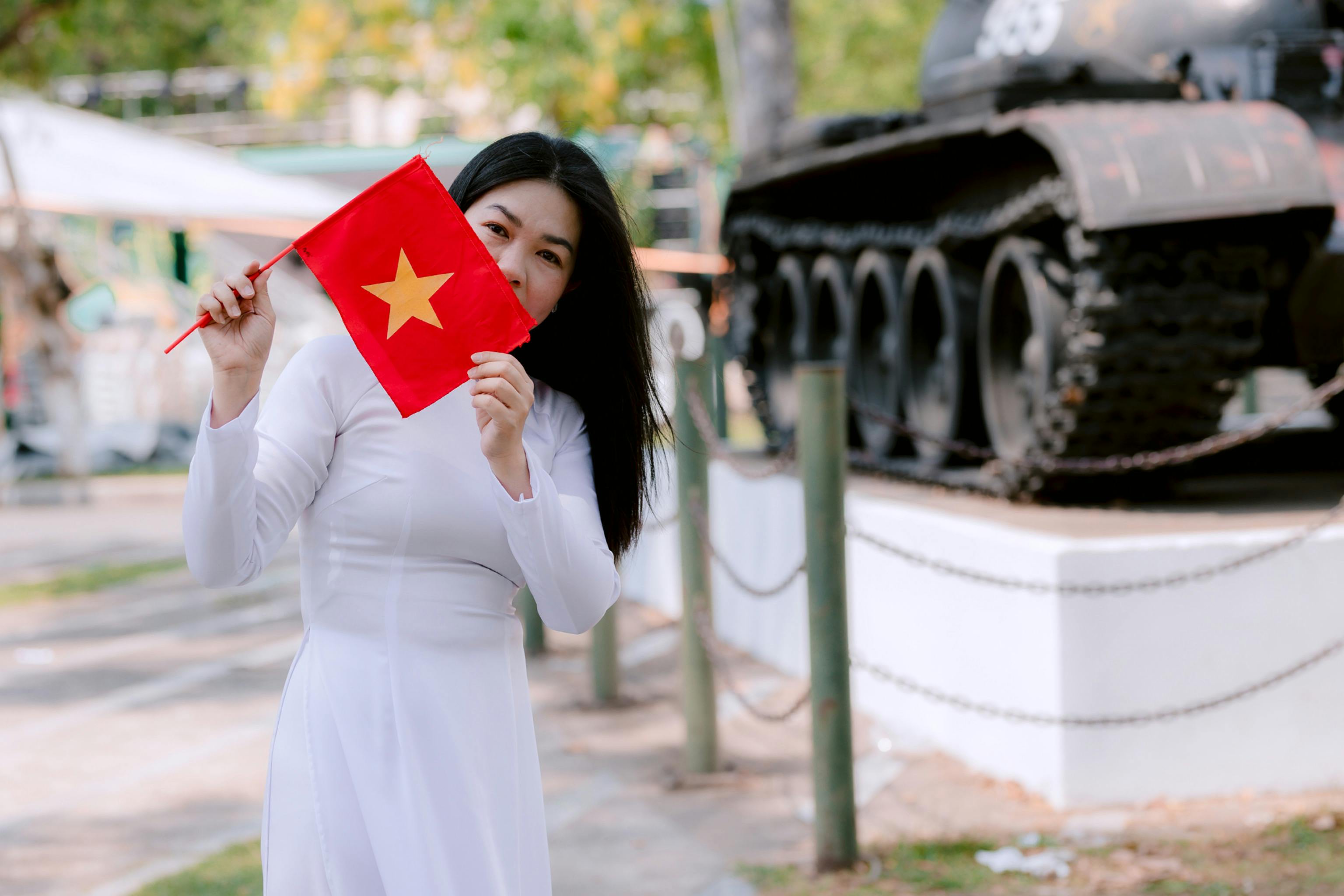 A woman in traditional dress holding the Vietnam flag near a historic war tank outdoors.